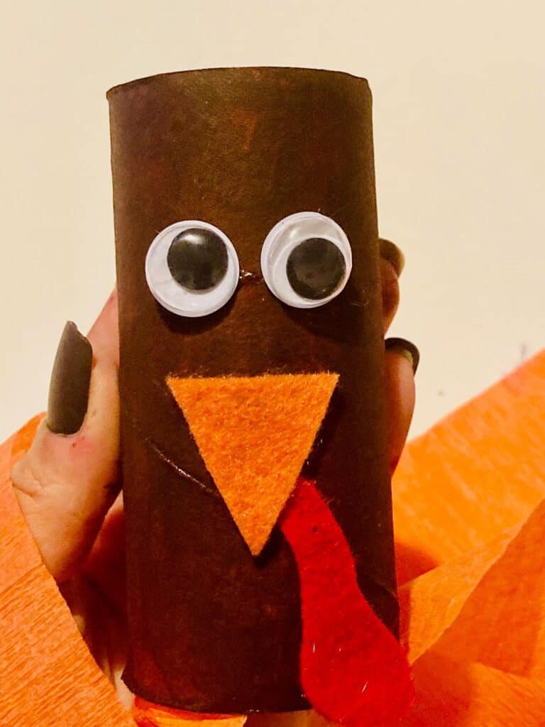Toilet paper roll turkey face with googly eyes and orange felt beak, and red snood glued to a brown painted empty toilet paper roll.