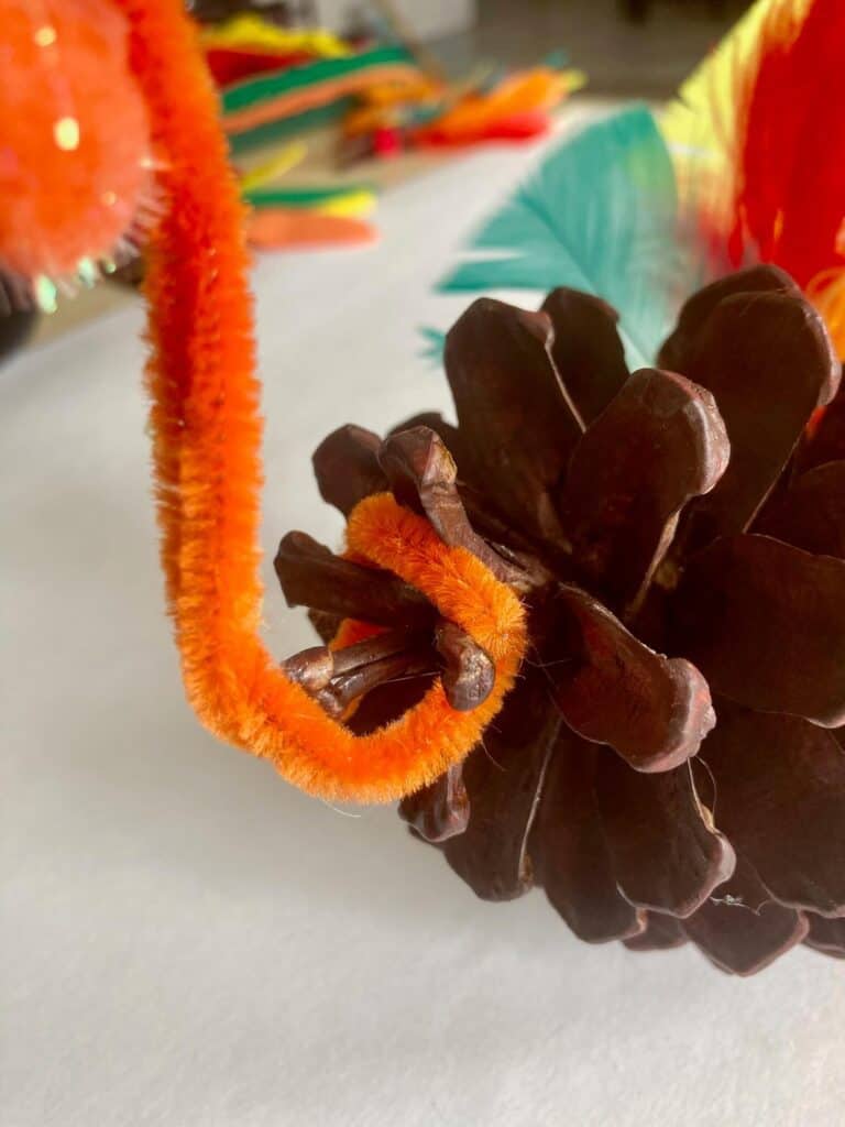 Orange pipe cleaner bent in the shape of a turkey neck, connected to the end of the brown painted pine cone.