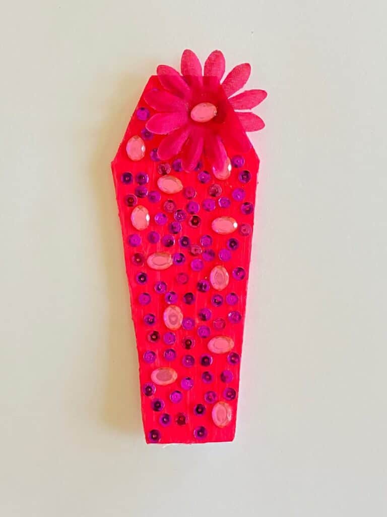 Pink painted foam board coffin with pink sequins and jewels as well as a pink flower embellishment.
