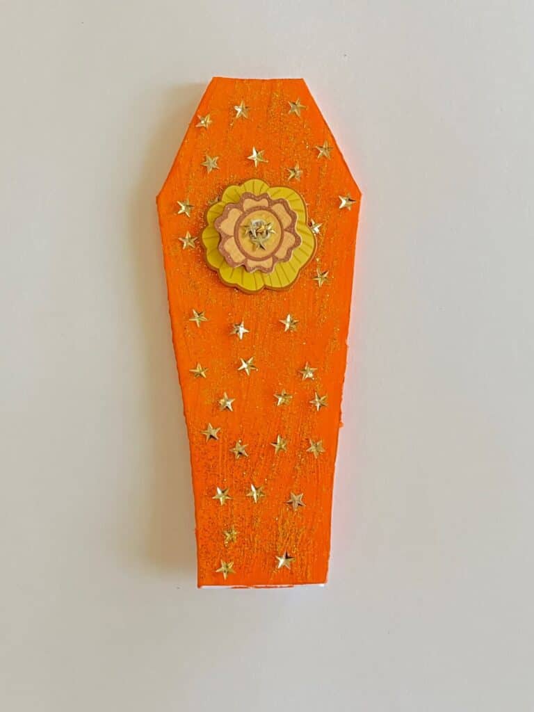 Orange painted foam board coffin with gold glitter and gold metallic stars. as well as a gold flower embellishment.