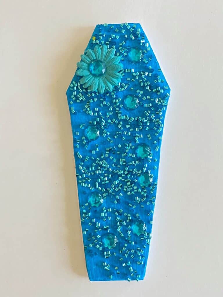 Blue painted foam board coffin with blue tube confetti and jewels as well as a blue flower embellishment.