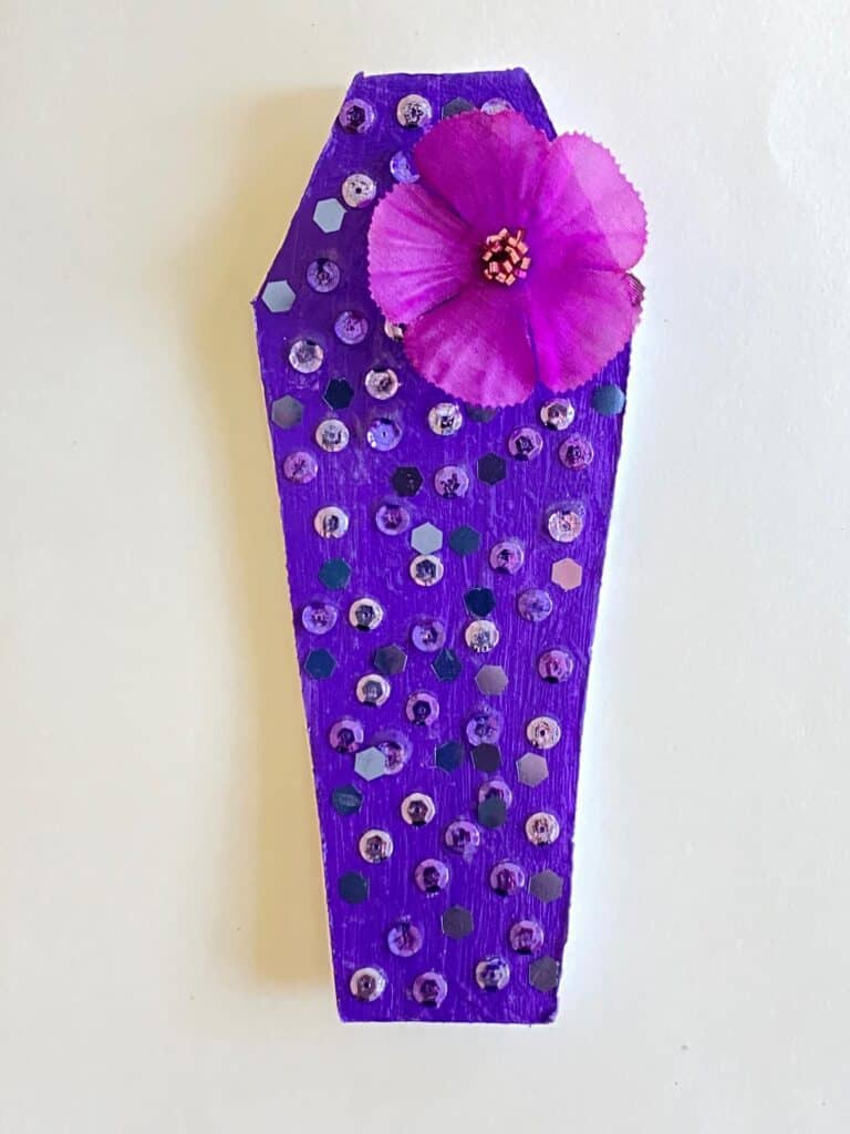Purple painted foam board coffin with purple sequins and glitter  as well as a purple flower embellishment.