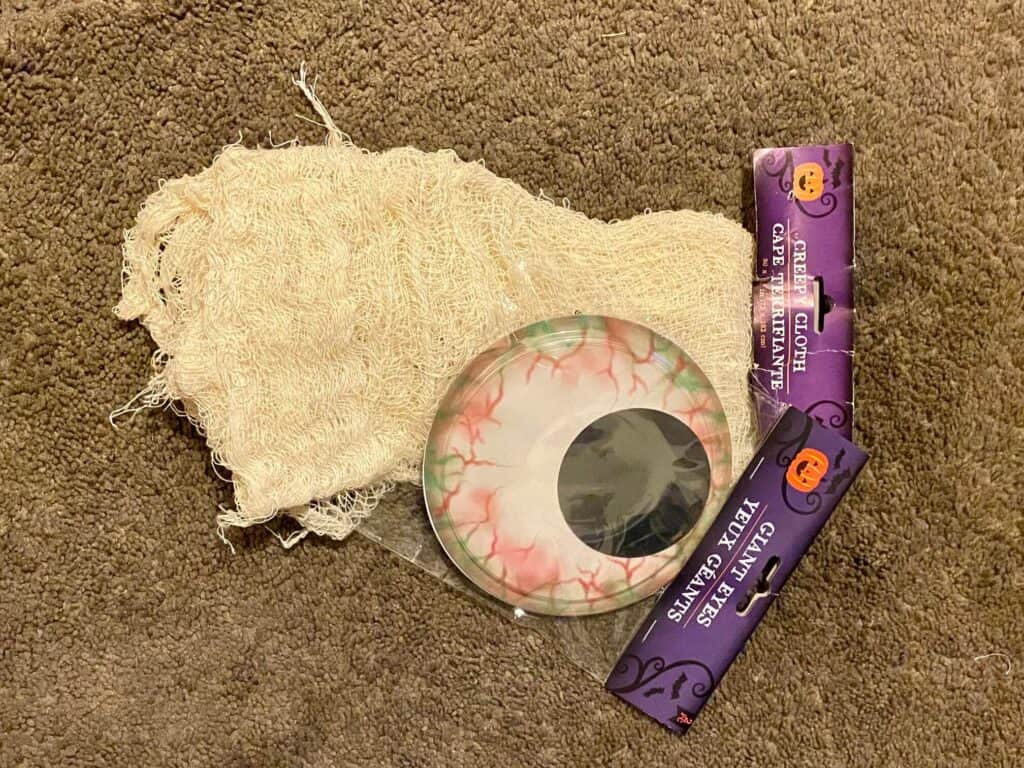 supplies needed to make a DIY Dollar Tree Mummy Door decoration laying on the floor is a white creepy cloth and jumbo bloodshot googly eyes.