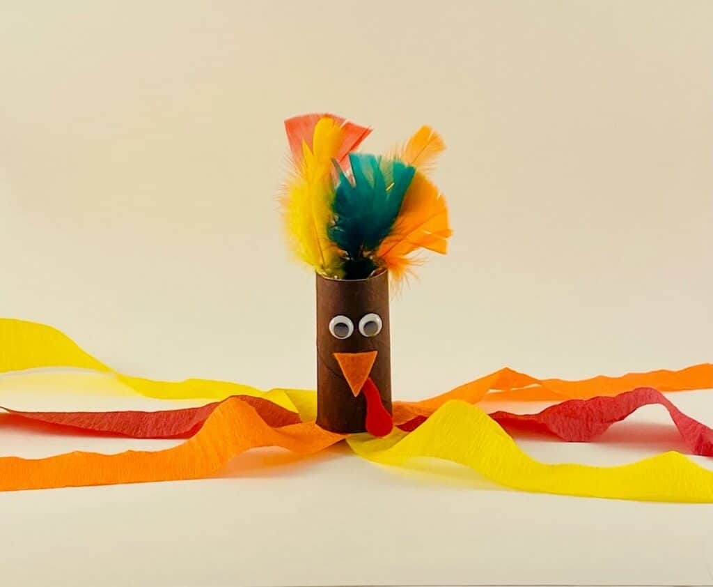 Toilet paper roll recycled craft Turkey windsock for kids, laying on the ground with the orange, yellow, and red crepe paper streamers spread out and feathers coming out of the turkey head.