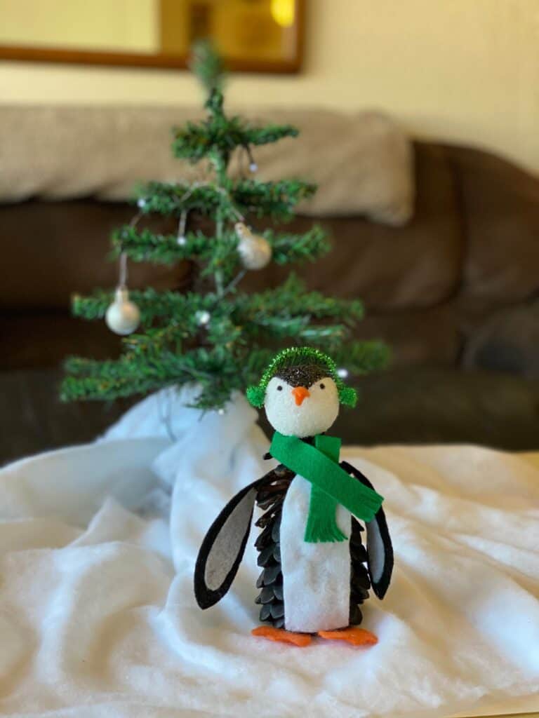 Pine Cone Penguin nature craft for kids with felt winter scarf and earmuffs and a Christmas tree behind him and fake snow underneath.