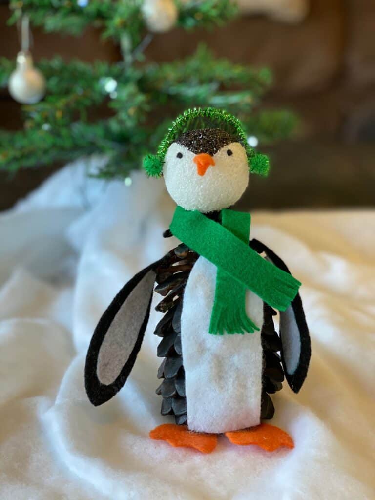 Pine Cone Penguin nature craft for kids with felt winter scarf and earmuffs and a Christmas tree behind him and fake snow underneath.