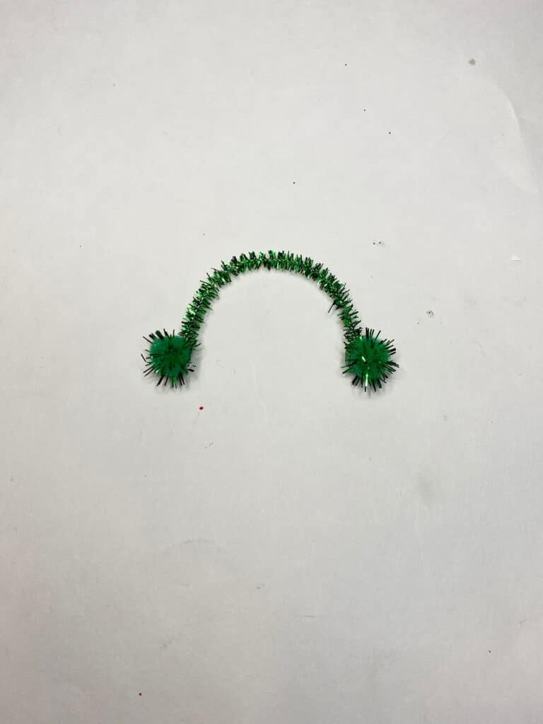 Small green piece of a pipe cleaner with a small green craft pom pom glued to each end to make earmuffs.