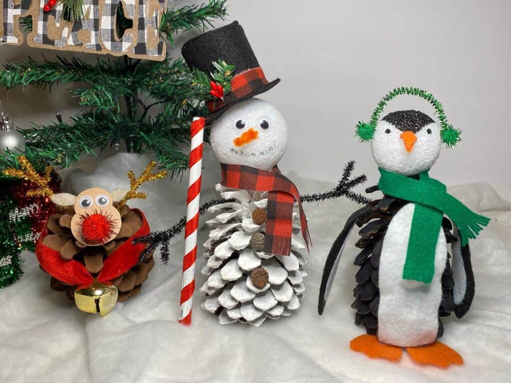 Pine cone reindeer, pine cone frosty the snowman, pine cone penguin all together with a christmas tree background and a snow blanket.