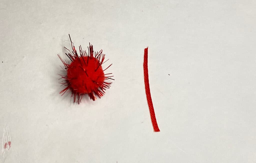 A red craft pom pom next to a small red piece of ribbon.