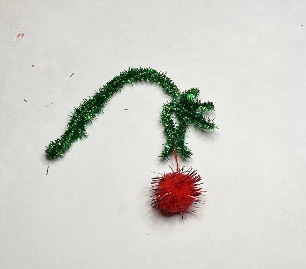 A green pipe cleaner shaped into the Grinch  hand holding a dangling red christmas bulb ornament.