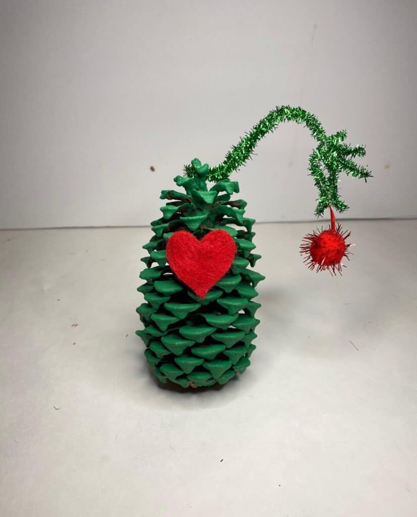 A pine cone painted green with a red felt heart on the front and a green pipe cleaner hand holding a dangling red christmas bulb ornament.