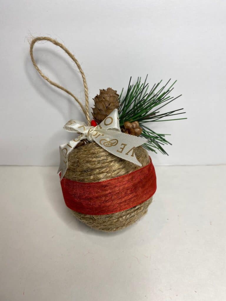 Handmade DIY Twine Wrapped Ball Christmas Tree ornament with a red sheer ribbon around the center, a white shoelace bow on top with a pine sprig, pine cone, and berry decoration.