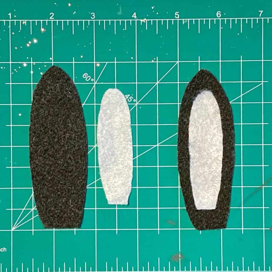 Black felt shaped pengiun wings with a white center, laying on a craft cutting mat.