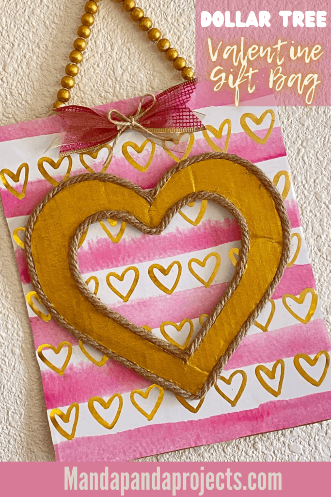 DIY Dollar Tree Pink and Gold Heart Valentine Gift Bag Sign with a gold wood bead hanger and a little bow hanging on the wall.