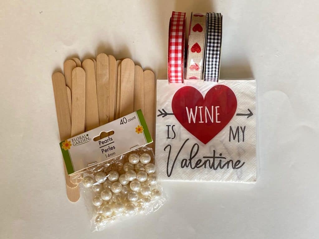 Supplies needed to make a DIY "Wine is my Valentine" napkin mod podged onto giant popsicle sticks with ribbon for bows and pearl bead hanger.