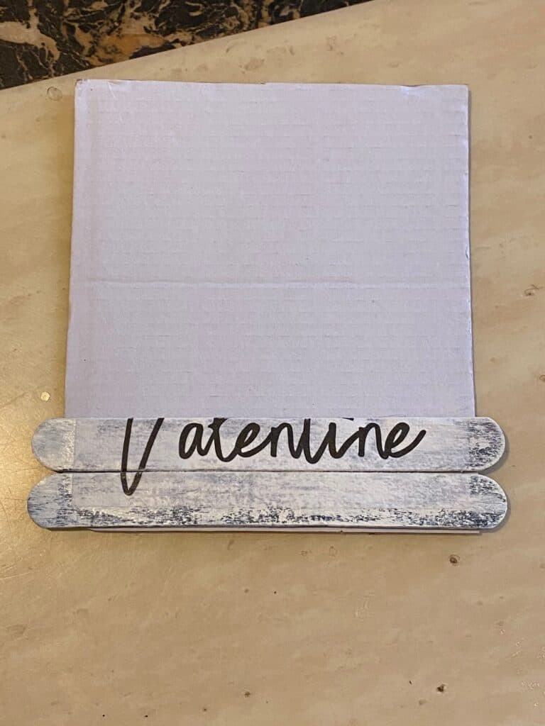 2 popsicle sticks glued to the bottom of a white cardboard square to make the Valentine sign.