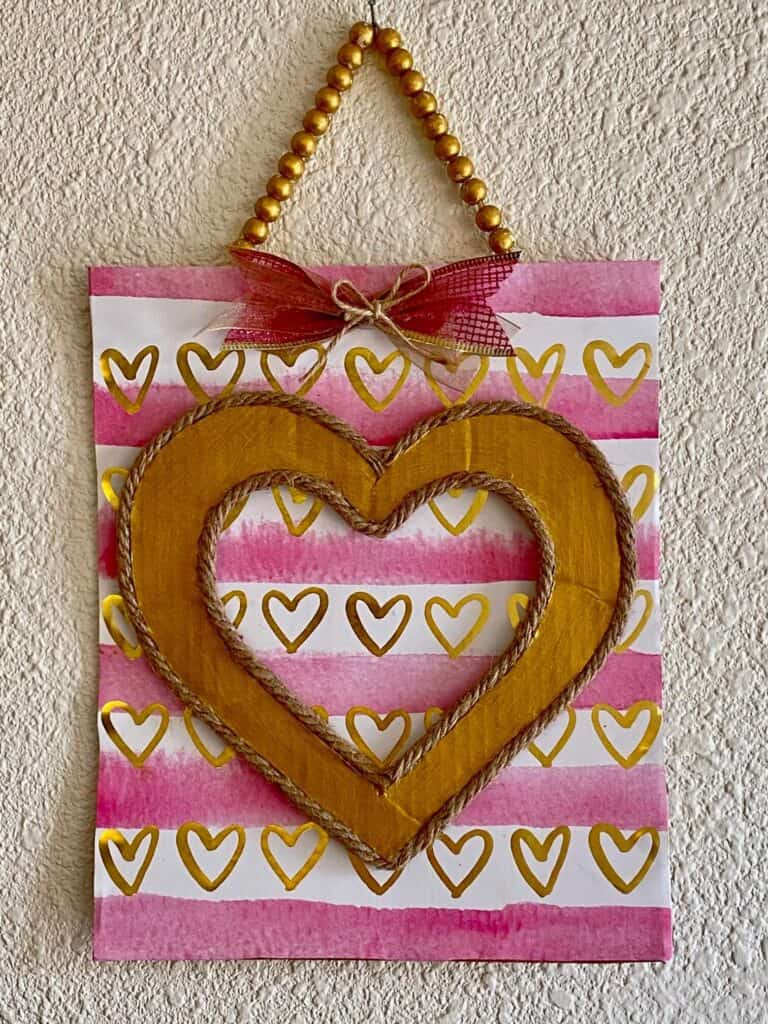 DIY Dollar Tree Pink and Gold Heart Valentine Gift Bag Sign with a gold wood bead hanger and a little bow hanging on the wall.
