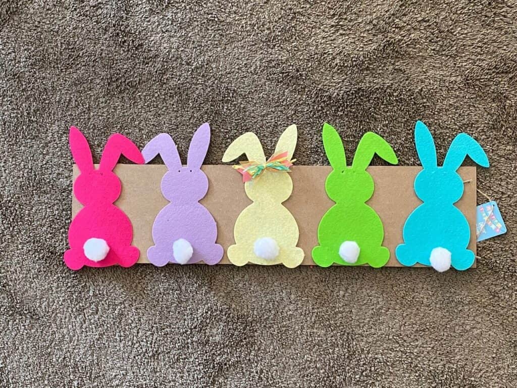 5 colored felt bunnies with cottonball tails on the back of a wooden rectangular sign.