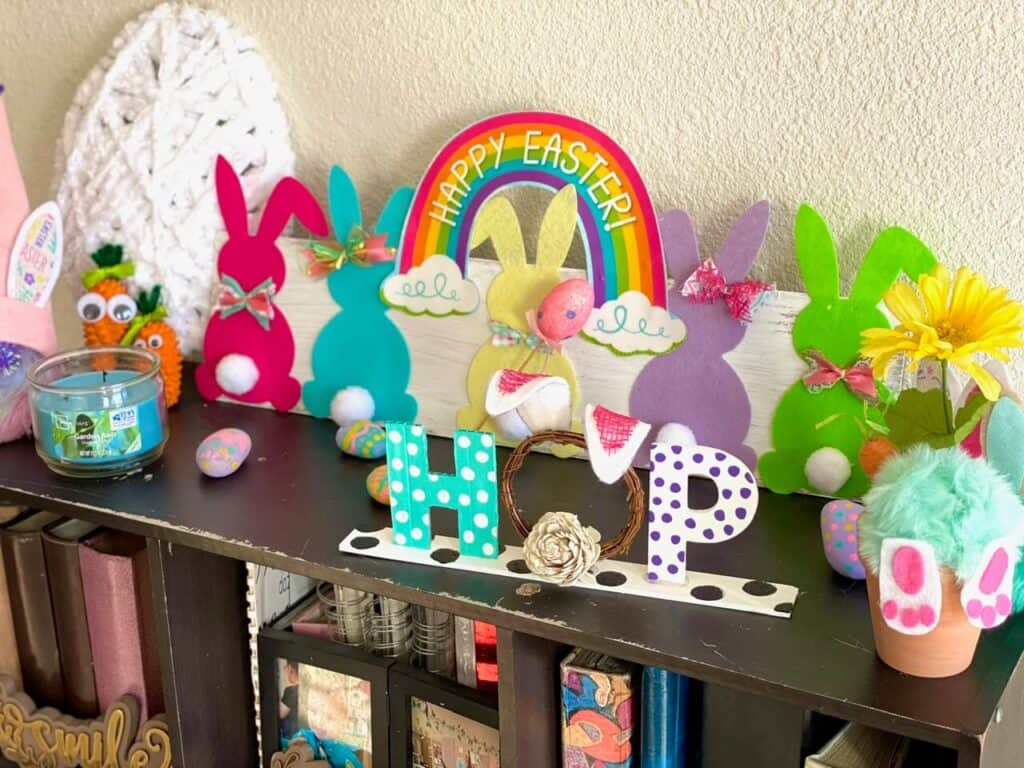 Top of a bookcase covered with super colorful Easter Holiday decor with the rainbow Bunny sign and a "hop" shelf sitter.