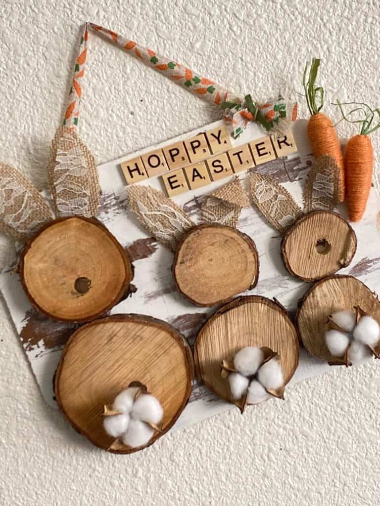 DIY rustic farmhouse wooden Easter bunny decor craft project with burlap ears, cotton pod tails, twine carrots, and 'Hoppy Easter' message in scrabble tiles, hanging with a carrot ribbon.