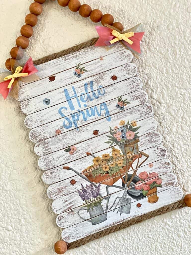 DIY Dollar Tree "Hello Spring" napkin sign made with popsicle stick planks for easy spring crafts and decor. Napkin has a wheelbarrow filled with sunflowers and garden tools.