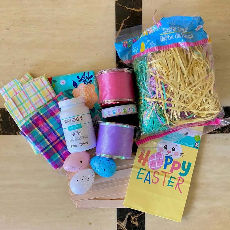 Supplies needed to make a dollar tree cutting board craft out of a Hoppy Easter gift bag, with fabric, ribbon, speckled eggs, colored easter grass, and chalk paint.