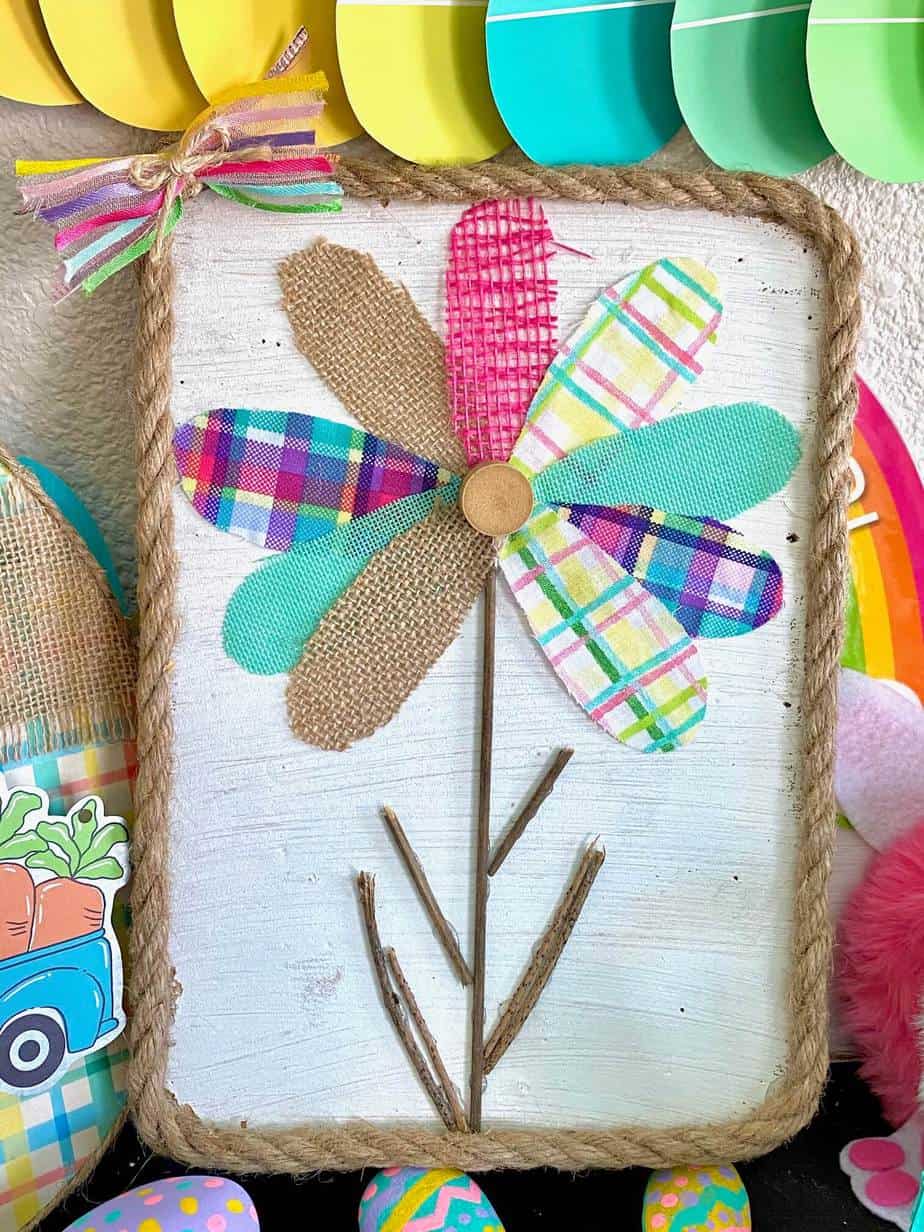 Scrap fabric and burlap ribbon colorful spring flower petals with a stick stem and white background with rope around the edge. DIY spring craft made with scraps.