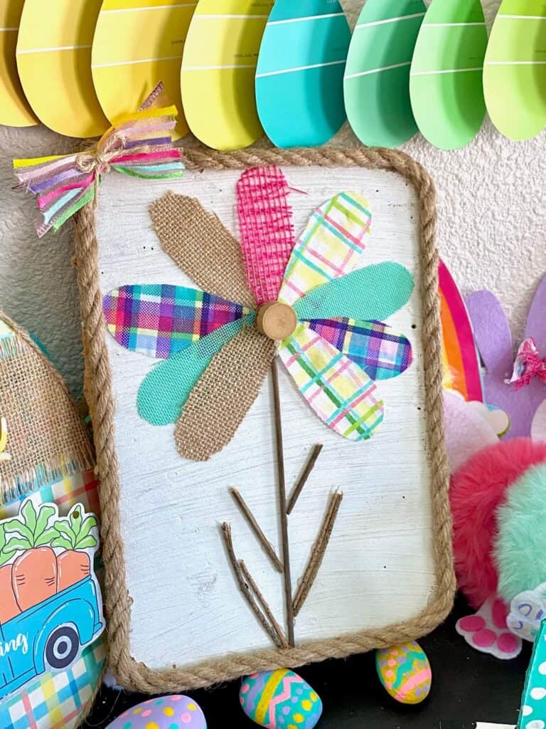 Scrap fabric and burlap ribbon colorful spring flower petals with a stick stem and white background with rope around the edge, sitting on a shelf underneath a colorful easter egg banner.