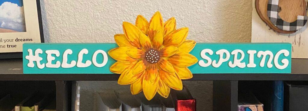 DIY Dollar Tree flower makeover with a Sunflower in the middle of a long teal colored wooden board with the word "Hello" on the left of the Sunflower and the word "Spring" on the right of the Sunflower.