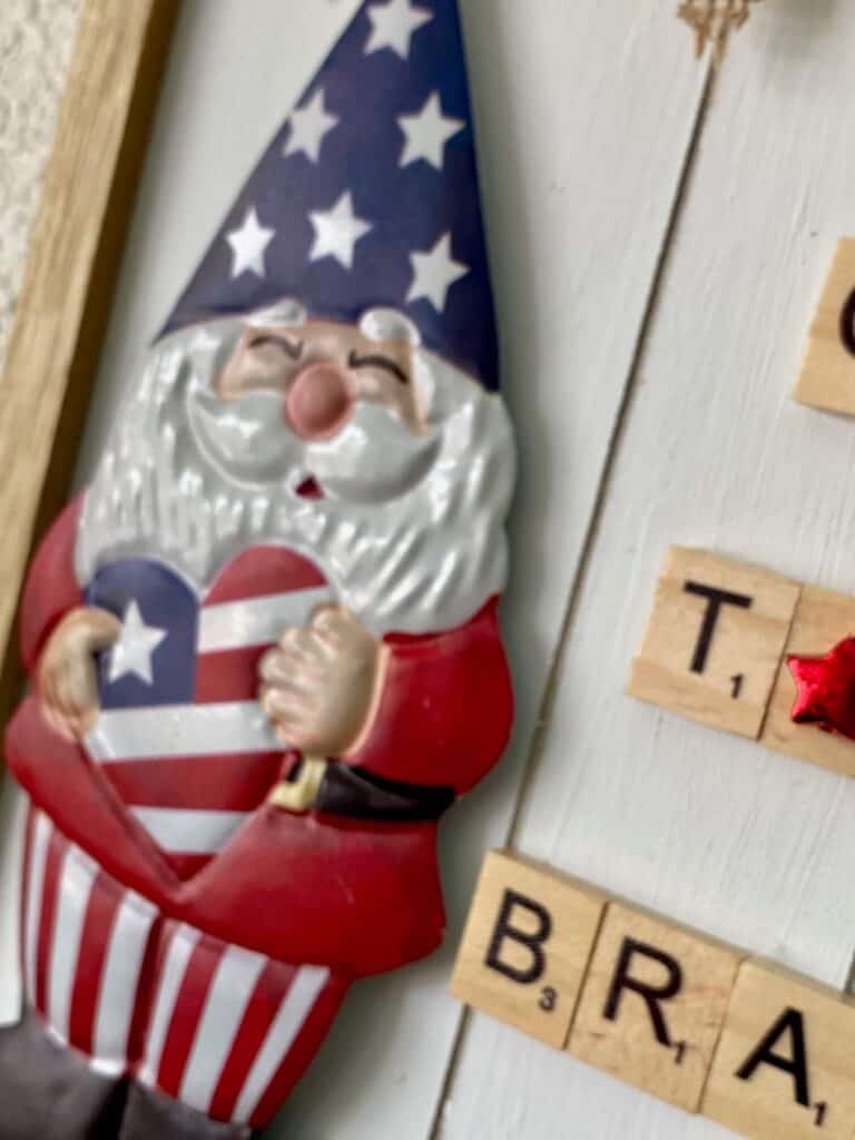 Close up of the Dollar Tree patriotic gnome DIY holding a red, white and blue heart and wearing stars and stripes, with some of the scrabble tiles visible.