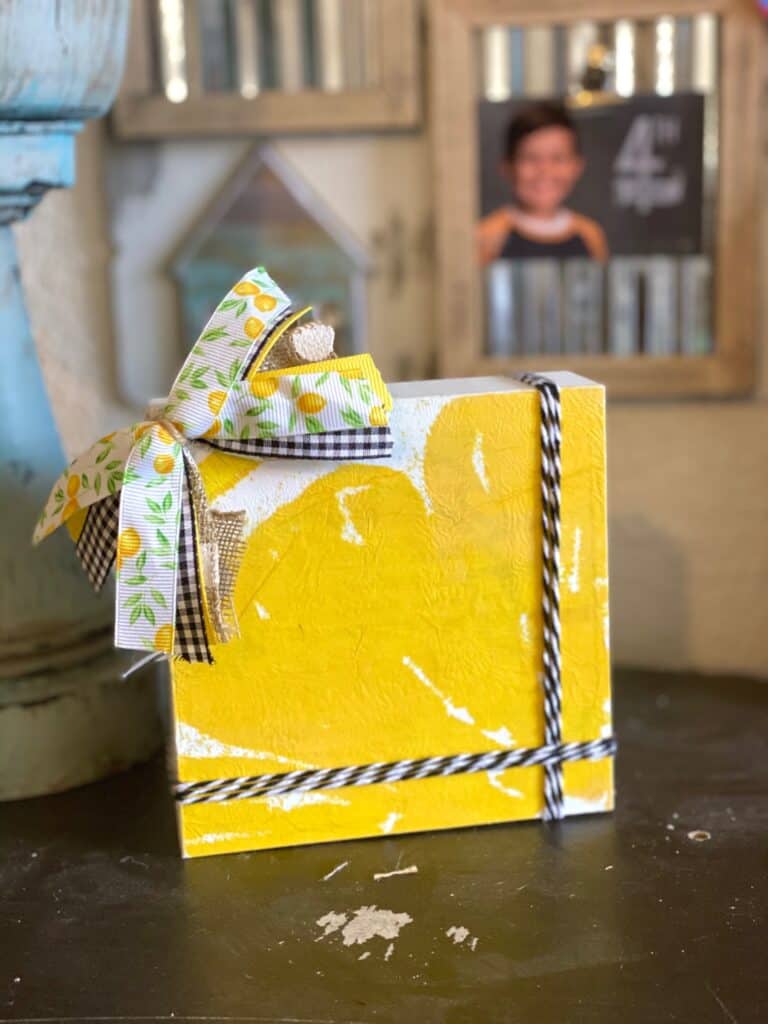 Dollar Tree Lemon Napkin Decoupaged onto a box frame to make a shelf sitter or DIY lemon tiered tray decor with black and white twine and a small lemon, burlap, and buffalo check bow in the top left corner.