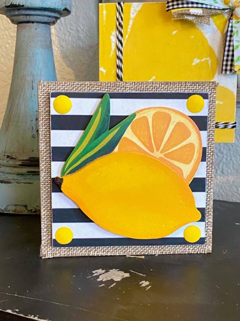 Lemon and lemon slice wooden cutout on a small wooden block with a black and white striped background on top of burlap with 4 yellow stickers in each corner.
