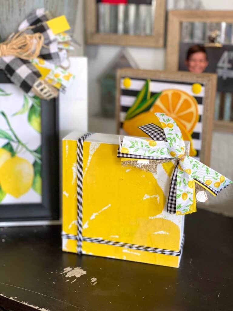 Dollar Tree Lemon Napkin Decoupaged onto a box frame to make a shelf sitter or DIY lemon tiered tray decor with black and white twine and a small lemon, burlap, and buffalo check bow in the top left corner.