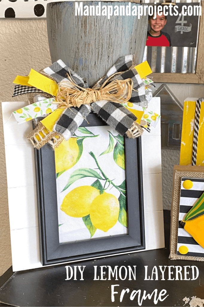 DIY summer Lemon Layered frame with a plain black dollar tree frame, Lemon fabric inside, layered on top of a white pallet board with a messy scrap ribbon bow on top, sitting on a bookshelf next to other lemon crafts and Decor.