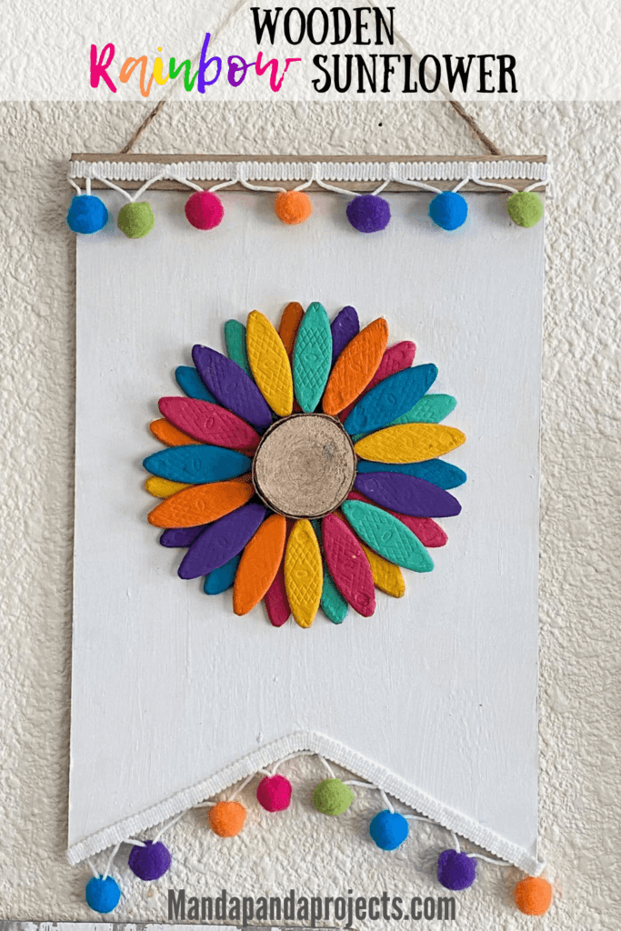 DIY Wooden rainbow sunflower made with wooden biscuits painted colorfully and a wooden round center, a white painted dollar tree background and rainbow pom pom trim along the top and bottom.