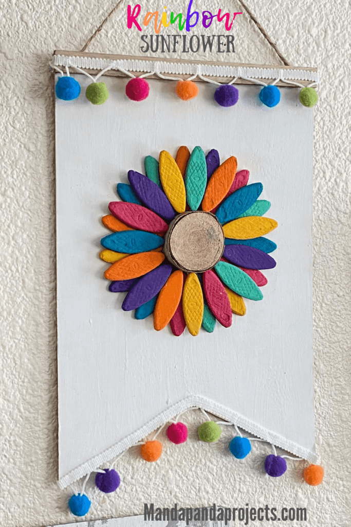 DIY Wooden rainbow sunflower made with wooden biscuits painted colorfully and a wooden round center, a white painted dollar tree background and rainbow pom pom trim along the top and bottom.