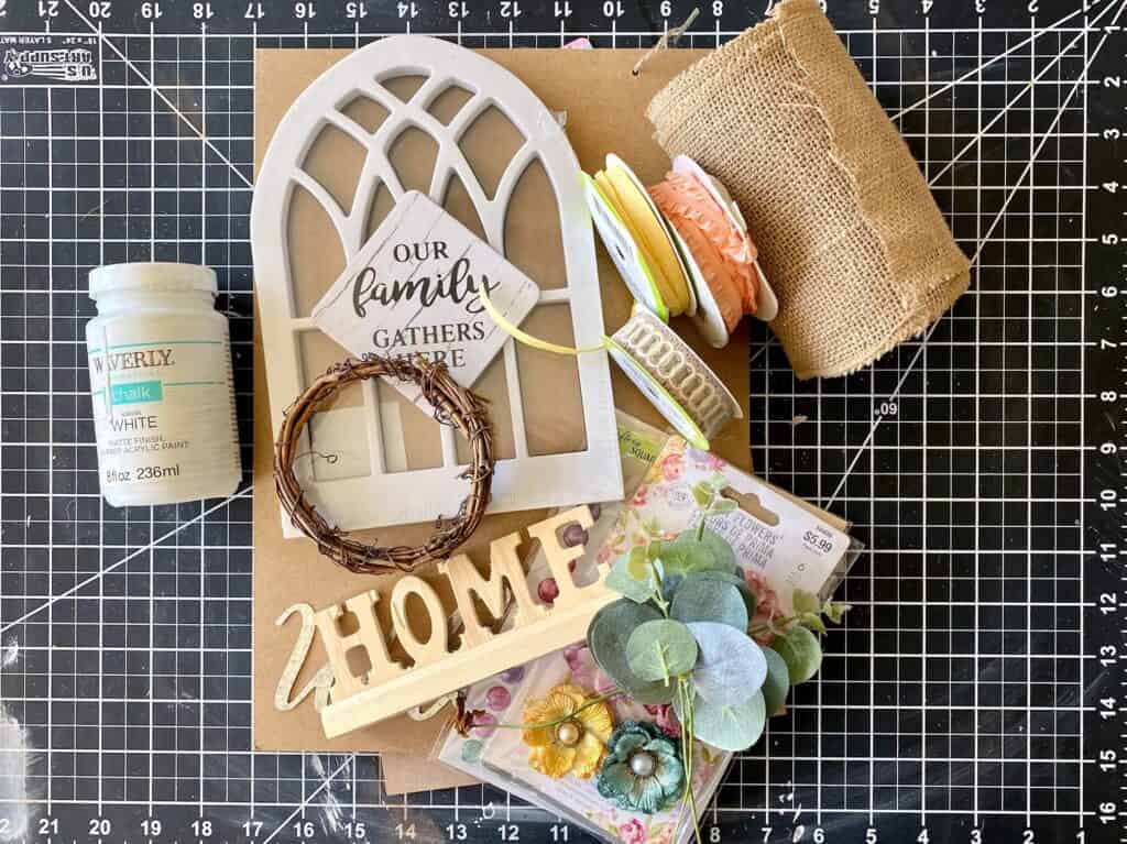 Supplies needed to make a dollar tree diy piece of decor with a cathedral window frame. Paint, burlap, metal welcome, flowers and grapevine wreath.