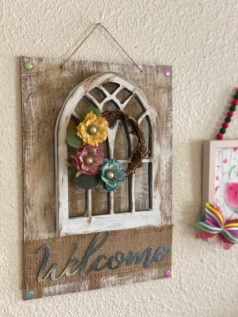 Side view of DIY Dollar Tree Cathedral window decor craft with a mini grapevine wreath and scrapbook embellishment flowers and a galvanized metal Welcome sign.