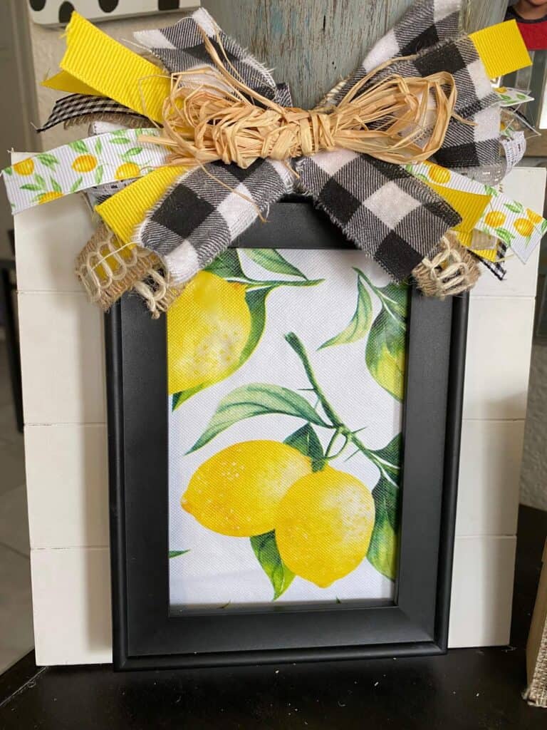 DIY summer Lemon Layered frame with a plain black dollar tree frame, Lemon fabric inside, layered on top of a white pallet board with a messy scrap ribbon bow on top.