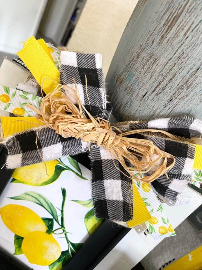 Scrap ribbon messy bow with raffia, buffalo check, lemon, yellow, and burlap ribbon on top of the layered frame.