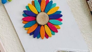 DIY Wooden Rainbow Sunflower with colorful flower petals on a dollar tree white painted background and pom pom trim.
