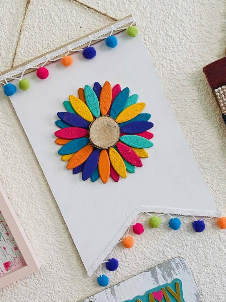 Sideways view of the DIY Wooden rainbow sunflower made with wooden biscuits painted colorfully and a wooden round center, a white painted dollar tree background and rainbow pom pom trim along the top and bottom.