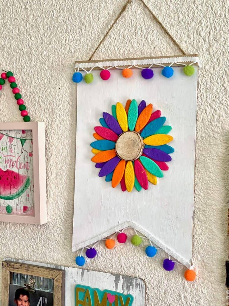 DIY wooden rainbow sunflower craft with rainbow painted flower petals on a wall next to a watermelon napkin frame.