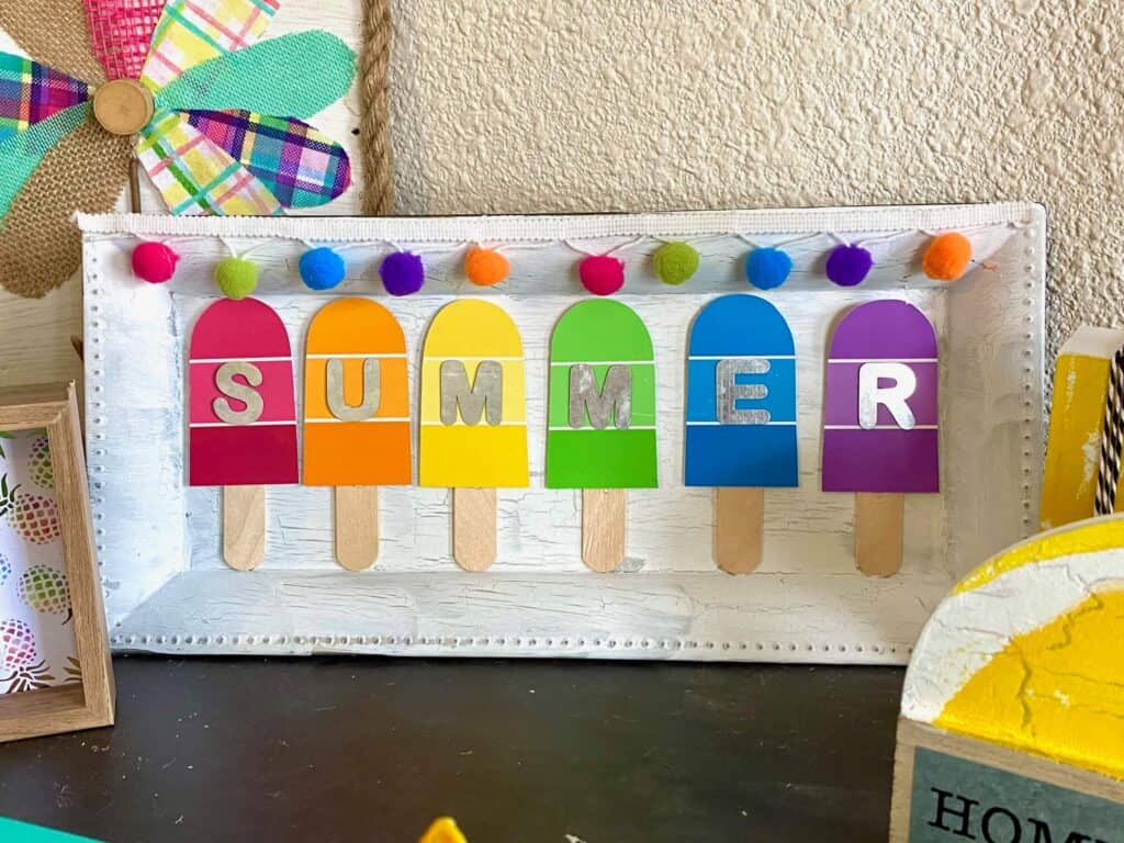 DIY Summer Paint Swatch popsicles on a dollar tree charger plate with rainbow pom pom trim on top.