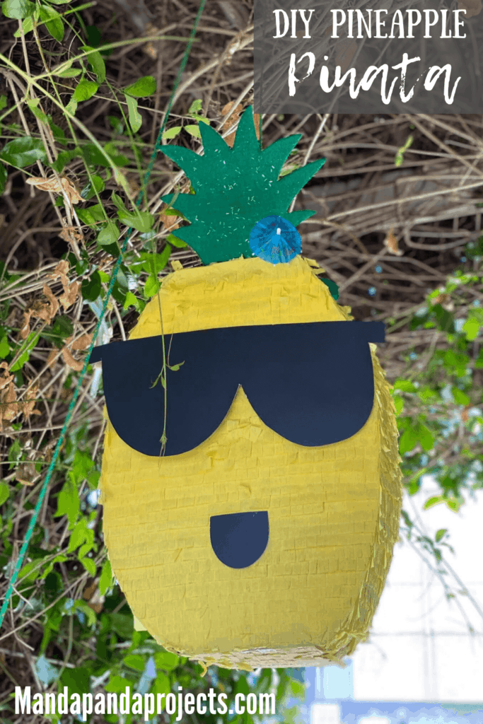 DIY Hawaiian Pineapple Piñata with a glitter foam crown, sunglasses, and mouth, hanging from an archway ready for the kids to hit it.