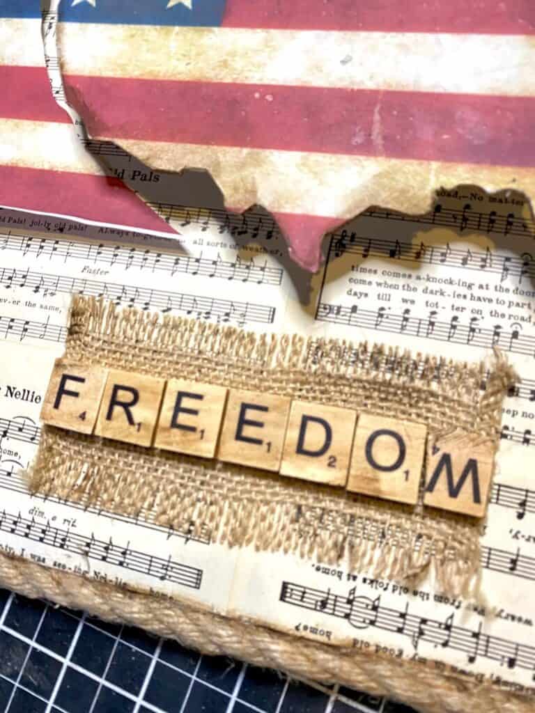 A strip of burlap with the word "freedom" spelled out in scrabble tile letters, underneath the American Flag USA cutout.