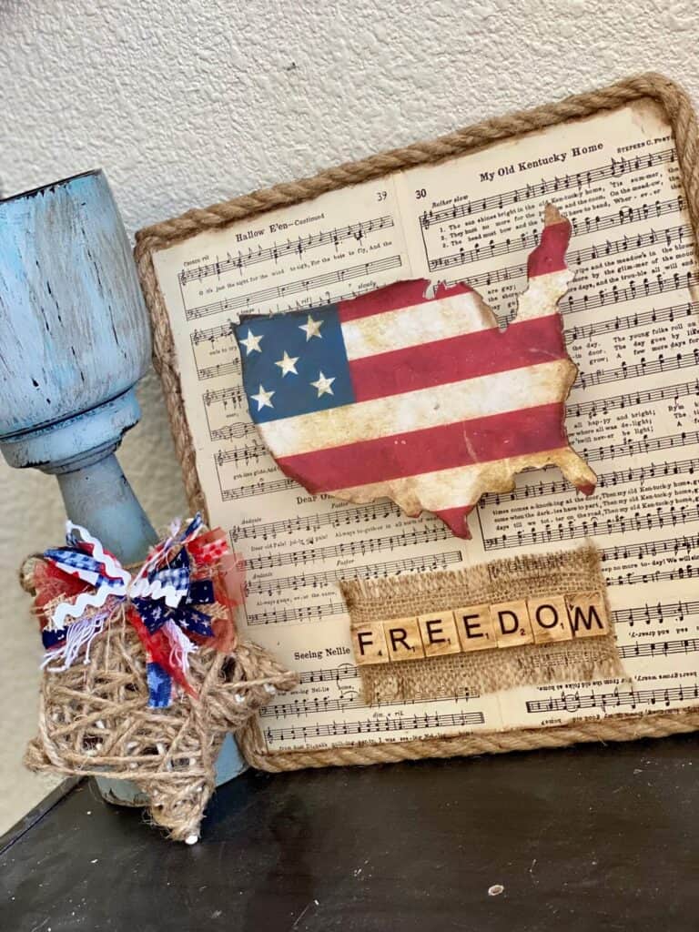 Dollar Tree DIY Fourth of July USA Freedom Flag Decor made with an America cutout and American flag paper on a music sheet background with nautical rope around the edge to celebrate the Americana Holiday.