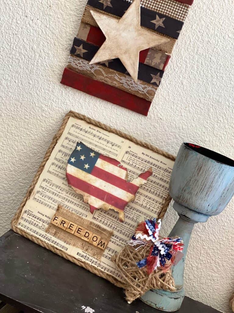 Dollar Tree DIY Fourth of July USA Freedom Flag Decor made with an America cutout and American flag paper on a music sheet background with nautical rope around the edge sitting next to a chunky candlestick and a rustic patriotic star hanging above.