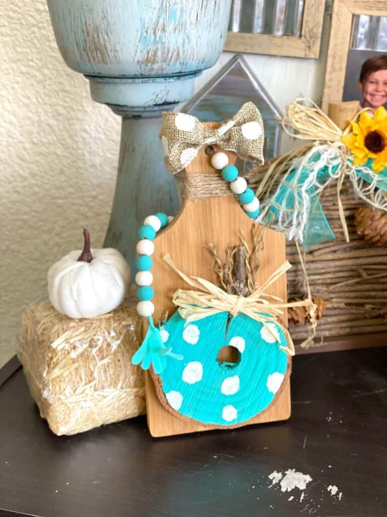 DIY Mini cutting board teal pumpkin fall tiered tray decor with a wood bead garland next to a hay bale and a white pumpkin.
