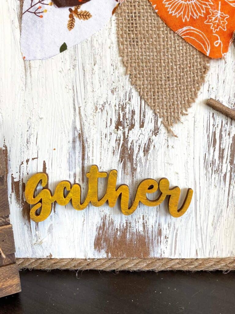 Close up of the wooden cutout word "gather" painted mustard yellow.